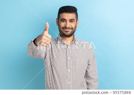 Portrait of businessman with beard standing with thumbs up, like gesture, demonstrating approval and agree with suggestion, wearing striped shirt. Indoor studio shot isolated on blue background. 95575869