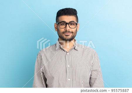Portrait of smiling positive pleased businessman in optical eyeglasses standing looking at camera with smile, wearing striped shirt. Indoor studio shot isolated on blue background. Portrait of smiling positive pleased businessman in optical eyeglasses standing looking at camera with smile, wearing striped shirt. Indoor studio shot isolated on blue background. 95575887