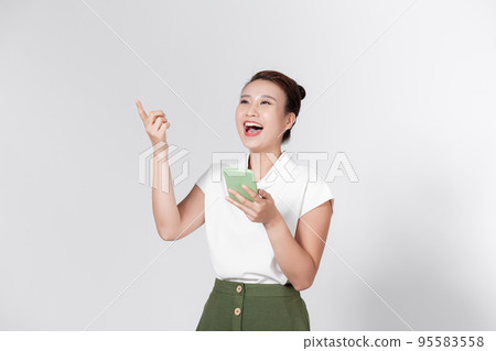 Overjoyed smiling happy cute Asian young lady point finger up hold phone posing isolated on white background. Overjoyed smiling happy cute Asian young lady point finger up hold phone posing isolated on white background. 95583558
