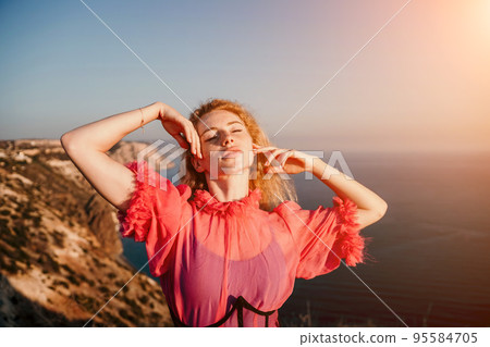 Close up portrait of curly redhead young caucasian woman with freckles looking at camera and smiling. Cute woman portrait in a pink long dress posing on a volcanic rock high above the sea at sunset Close up portrait of curly redhead young caucasian woman with freckles looking at camera and smiling. Cute woman portrait in a pink long dress posing on a volcanic rock high above the sea at sunset 95584705