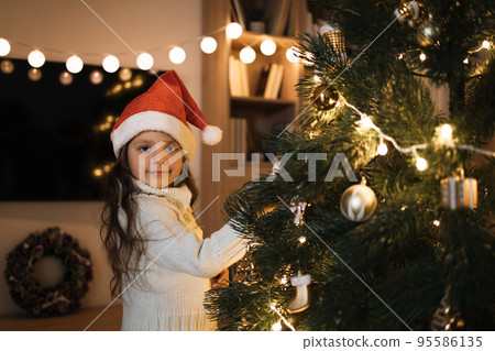 Little adorable girl in knitted sweater is hanging decoration on Christmas tree with light. Little adorable girl in knitted sweater is hanging decoration on Christmas tree with light. 95586135