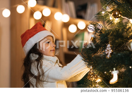 Little adorable girl in knitted sweater is hanging decoration on Christmas tree with light. Little adorable girl in knitted sweater is hanging decoration on Christmas tree with light. 95586136
