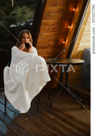 Pretty woman sips tea while sitting on the terrace of bungalow wrapped in a blanket Pretty woman sips tea while sitting on the terrace of bungalow wrapped in a blanket 95587790