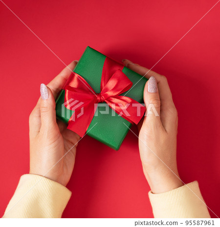 Woman hands holding Christmas present. Gift box wrapped in green paper with red ribbon. 95587961