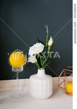 A white ceramic vase with an artificial white eustoma flower in it and a lemon in a stemmed glass. Decor in the dining room or kitchen 95588934