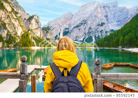Backview of the woman with a backpack and wearing a yellow sport jacket having fun on lake Braies with turquoise water and wooden boats in the Dolomites. 95592145