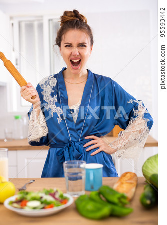 Angry woman screaming and waving rolling pin in home kitchen Angry woman screaming and waving rolling pin in home kitchen 95593442