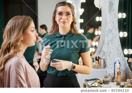 Female beauty specialist in glasses looking at camera and smiling while woman sitting at dressing table in beauty salon or visage studio. Makeup artist holding cosmetic brush and eyebrow shadow. Female beauty specialist in glasses looking at camera and smiling while woman sitting at dressing table in beauty salon or visage studio. Makeup artist holding cosmetic brush and eyebrow shadow. 95594210