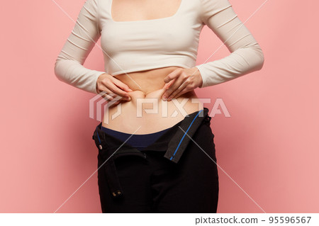Closeup female hands stretching skin on her belly, model showing to camera her abdomen. Concept of dieting, sport, fitness, healthy lifestyle, body positive and wellbeing Closeup female hands stretching skin on her belly, model showing to camera her abdomen. Concept of dieting, sport, fitness, healthy lifestyle, body positive and wellbeing 95596567