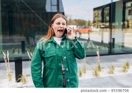 Beautiful Bored redhead woman talking with her smartphone. Woman yawning and using smartphone on city street. Beautiful Bored redhead woman talking with her smartphone. Woman yawning and using smartphone on city street. 95596706