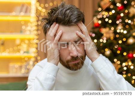 A man has a headache for Christmas, a close-up photo on New Year's Eve near the tree, a man holding his head with his hands, sick, sitting on the sofa at home alone. 95597070