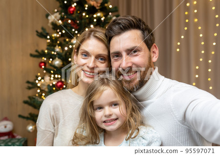 Portrait of happy family on Christmas, family man woman and daughter looking at camera and smiling, celebrating new year together at home, using phone for video call and remote communication online. 95597071