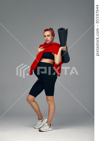 Pregnant woman holding yoga mat in hands, going for sport while awaiting baby, looking smiling at camera and touching her belly, standing against gray wall. Pregnant woman holding yoga mat in hands, going for sport while awaiting baby, looking smiling at camera and touching her belly, standing against gray wall. 95597644