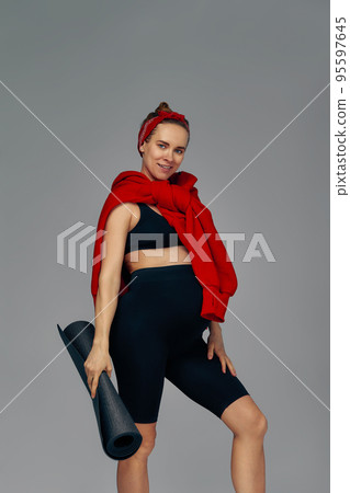 Pregnant woman holding yoga mat in hands, going for sport while awaiting baby, looking smiling at camera and touching her belly, standing against gray wall. Pregnant woman holding yoga mat in hands, going for sport while awaiting baby, looking smiling at camera and touching her belly, standing against gray wall. 95597645