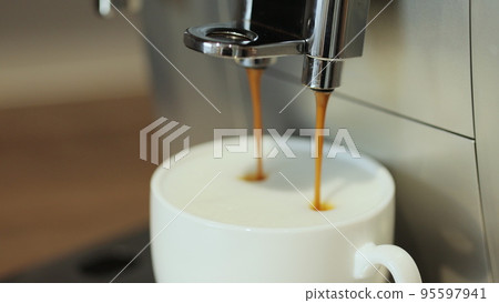 Making cappuccino using automatic coffee machine. Coffee is pouring into cup with frothed milk or cream close-up. Beverage drink for breakfast. Making cappuccino using automatic coffee machine. Coffee is pouring into cup with frothed milk or cream close-up. Beverage drink for breakfast. 95597941