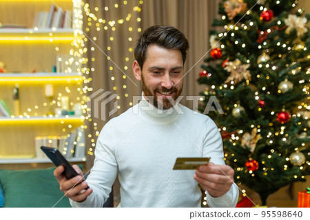 Man alone at home on Christmas celebrating new year, sitting on sofa and holding smartphone and bank credit card, happy smiling online shopping choosing gifts for friends and family remotely. 95598640
