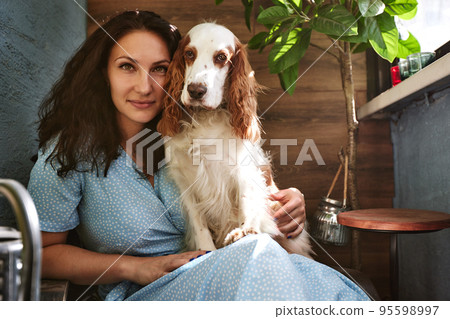 Cheerful young woman holding her big puppy with black nose and laughing. Indoor portrait of smiling girl with dark short hair posing with dog on background at home. Cheerful young woman holding her big puppy with black nose and laughing. Indoor portrait of smiling girl with dark short hair posing with dog on background at home. 95598997