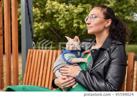 The young woman is holding her cat in amoveralls on her feet. Cat on a walk. Love to the animals. Close-up. 95599024