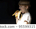 Funny portrait of boy with blond hair eating banana on black background. 95599315