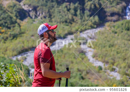 A hiker with canes looks into the distance on a path in the mountains to the waterfalls. 95601826