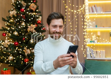 Cheerful man at home uses the phone, celebrates Christmas and New Year sitting on the sofa near the Christmas tree, types messages and browses online on the Internet. 95602851