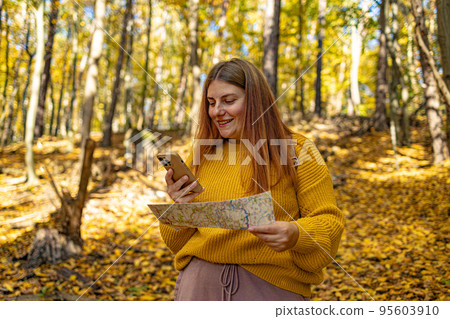Attractive hiker girl walking along forest path in mountains and using smartphone, looking away. Hiker woman in red raincoat navigates on smartphone on a hike. Use the map when traveling. 95603910