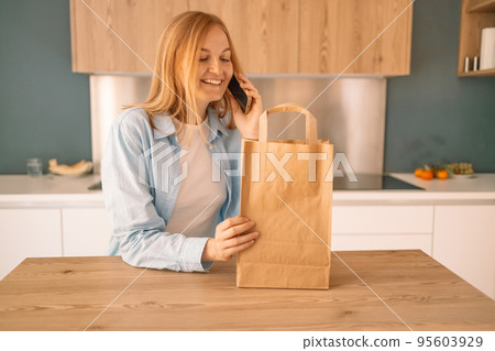 Beautiful young caucasian woman hold eco shopping bag after shopping using smartphone in modern kitchen at home Beautiful young caucasian woman hold eco shopping bag after shopping using smartphone in modern kitchen at home 95603929
