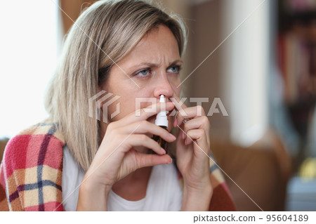 Ill woman sitting on sofa wrapped in blanket using nose drops Ill woman sitting on sofa wrapped in blanket using nose drops 95604189