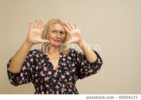 Pretty blonde woman with long hair posing in dark dress on a beige background. Emotional portrait. She makes a scared face and holds her hands in front of her to protect herself from her 95604225