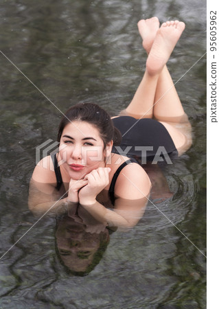Overweight young woman in black one-piece bathing suit lying down, relaxation in geothermal mineral water in outdoors pool balneotherapy health spa, hot springs resort having balneological properties 95605962