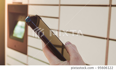 Female hand holding mobile phone next to Parcel locker Collecting parcel from shopping locker. Woman Skans QR Code on Mobile phone Self-service Locker Cell with Bar code Reader Modern Shipping and 95606122