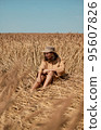 Young woman barefoot in linen clothes and a hat on a background of dry grass. Romantic girl in a hat sits on a meadow, relaxed alone in nature. 95607826