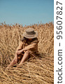 Young woman barefoot in linen clothes and a hat on a background of dry grass. Romantic girl in a hat sits on a meadow, relaxed alone in nature. 95607827