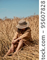 Young woman barefoot in linen clothes and a hat on a background of dry grass. Romantic girl in a hat sits on a meadow, relaxed alone in nature. 95607828