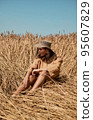 Young woman barefoot in linen clothes and a hat on a background of dry grass. Romantic girl in a hat sits on a meadow, relaxed alone in nature. 95607829