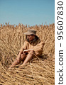 Young woman barefoot in linen clothes and a hat on a background of dry grass. Romantic girl in a hat sits on a meadow, relaxed alone in nature. 95607830