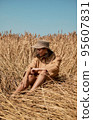 Young woman barefoot in linen clothes and a hat on a background of dry grass. Romantic girl in a hat sits on a meadow, relaxed alone in nature. 95607831