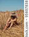 Young woman barefoot in linen clothes and a hat on a background of dry grass. Romantic girl in a hat sits on a meadow, relaxed alone in nature. 95607838