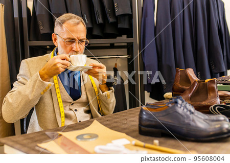 An elderly shoemaker taking a break from sewing with cup of coffee in a workshop 95608004