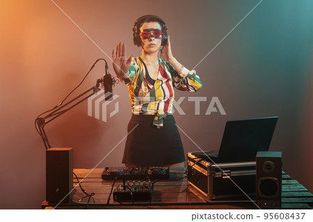 Young artist doing stop sign with palm and listening to sounds in headphones, mixing music at turntables. Cool female DJ showing rejection and denial gesture, forbidden or refusal. 95608437