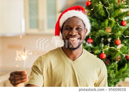 african american man in red Santa hat holding sparkler near christmas tree at home 95609038