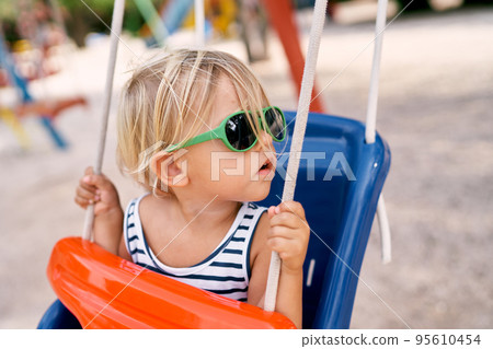 Little girl sits on a swing holding the ropes with her hands and turning her head. High quality photo 95610454