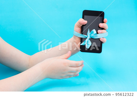Digital detox concept. A children's hand with a smartphone is tied with a blue ribbon and a second hand reaches for them. Gadget addiction Digital detox concept. A children's hand with a smartphone is tied with a blue ribbon and a second hand reaches for them. Gadget addiction 95612322
