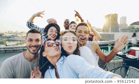 Point of view shot of happy friends taking selfe on roof at summer party laughing, posing and enjoying good company. Happiness, leisure and modern technology concept. Point of view shot of happy friends taking selfe on roof at summer party laughing, posing and enjoying good company. Happiness, leisure and modern technology concept. 95612542
