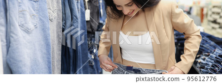 Young woman shopaholic choosing jeans in luxury store, woman try on pants on big sale in shop Young woman shopaholic choosing jeans in luxury store, woman try on pants on big sale in shop 95612776