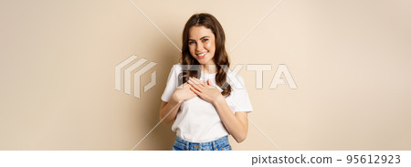 Portrait of young caucasian woman hold hands on heart and smiling, daydreaming, thinking of smth heartwarming, posing against beige background Portrait of young caucasian woman hold hands on heart and smiling, daydreaming, thinking of smth heartwarming, posing against beige background 95612923