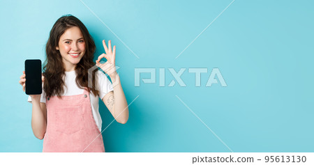 Portrait of modern stylish girl recommending online shop or mobile app, showing okay sign with empty smartphone screen, nod in approval, smiling satisfied, blue background 95613130