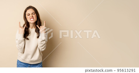 Happy young woman enjoying music, having fun, showing rock on, heavy metal finger horns gesture, standing over beige background 95613291