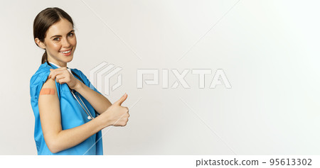 Smiling nurse, medical worker showing thumbs up and shoulder after vaccine, covid-19 vaccination campaign, standing over white background in scrubs Smiling nurse, medical worker showing thumbs up and shoulder after vaccine, covid-19 vaccination campaign, standing over white background in scrubs 95613302