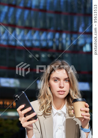Stylish young corporate woman standing in street, drinks coffee and holds mobile phone 95613513